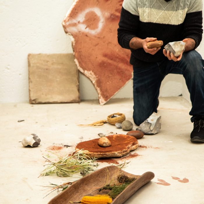 Man with two rocks kneeling on concrete floor with Aboriginal artifacts in front